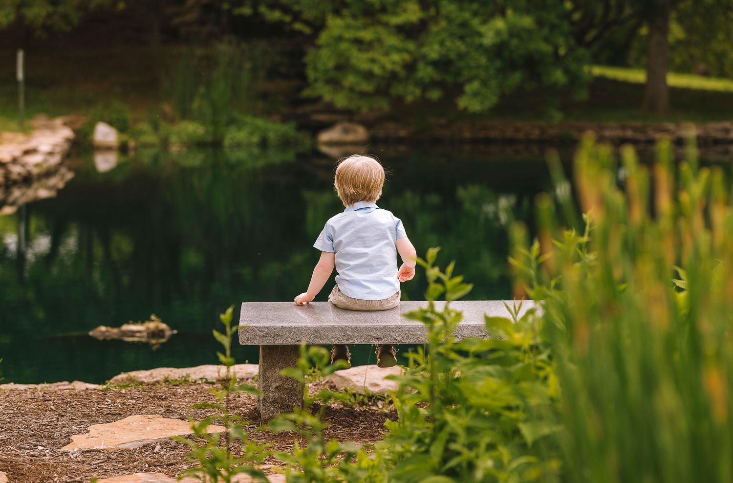 A small child in white sits alone on a wooden bench overlooking a serene pond surrounded by lush greenery.
