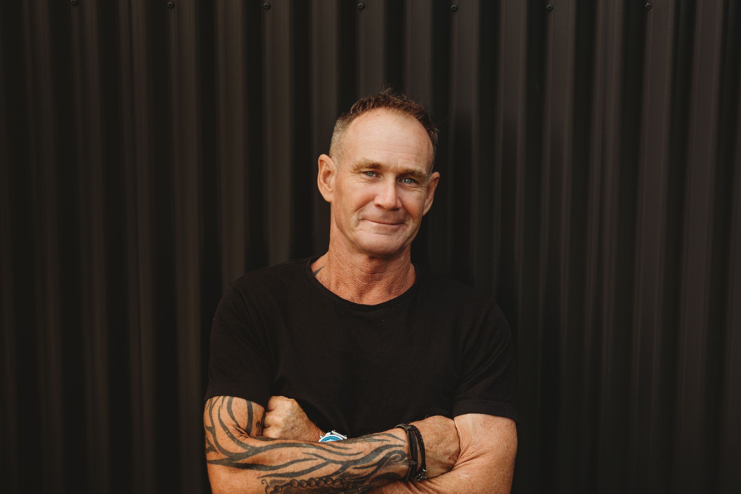 Person in black t-shirt smiling against dark background showing tattoo sleeve in casual portrait.