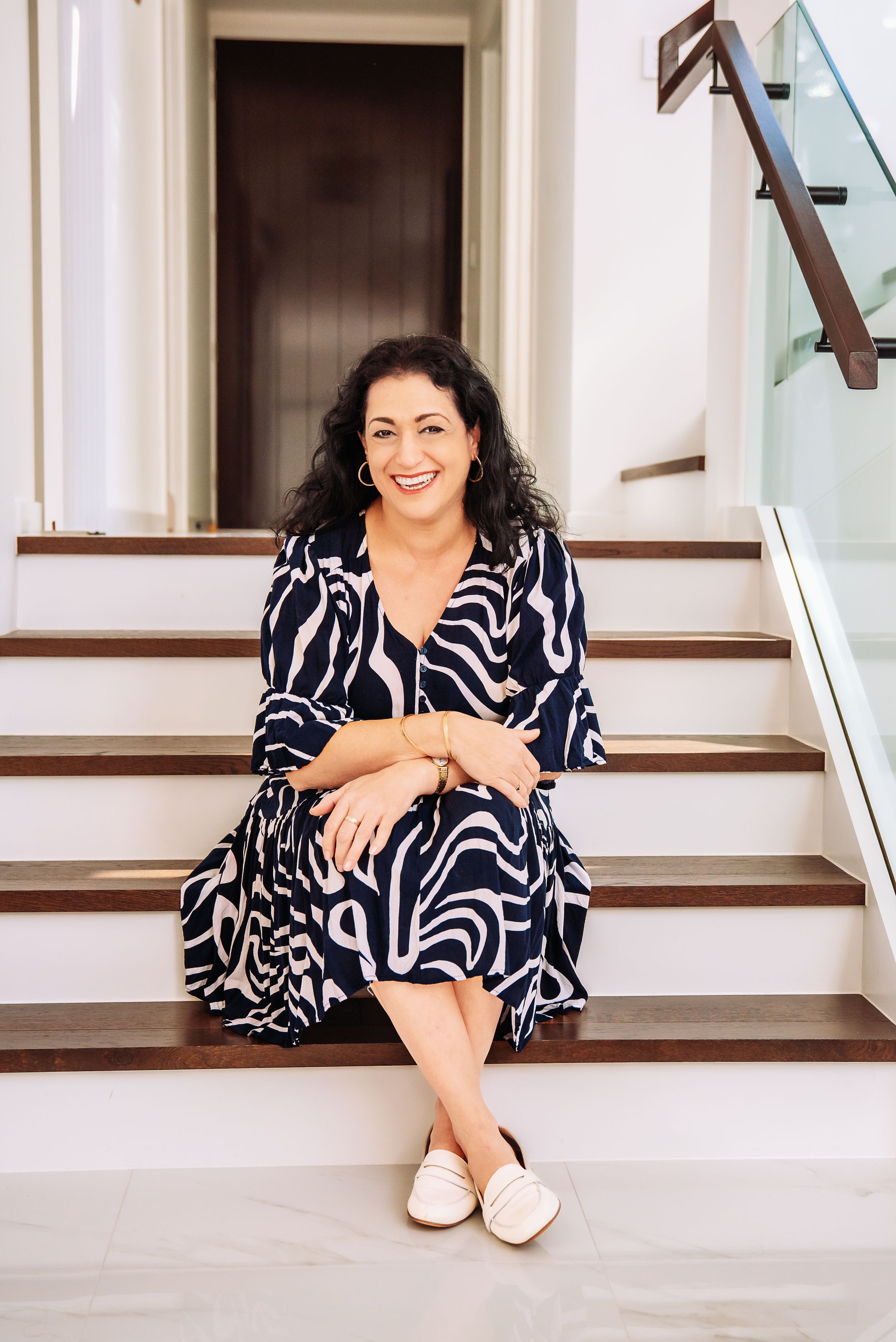 A person in a flowy black and white patterned dress sits casually on wooden stairs near a glass railing.