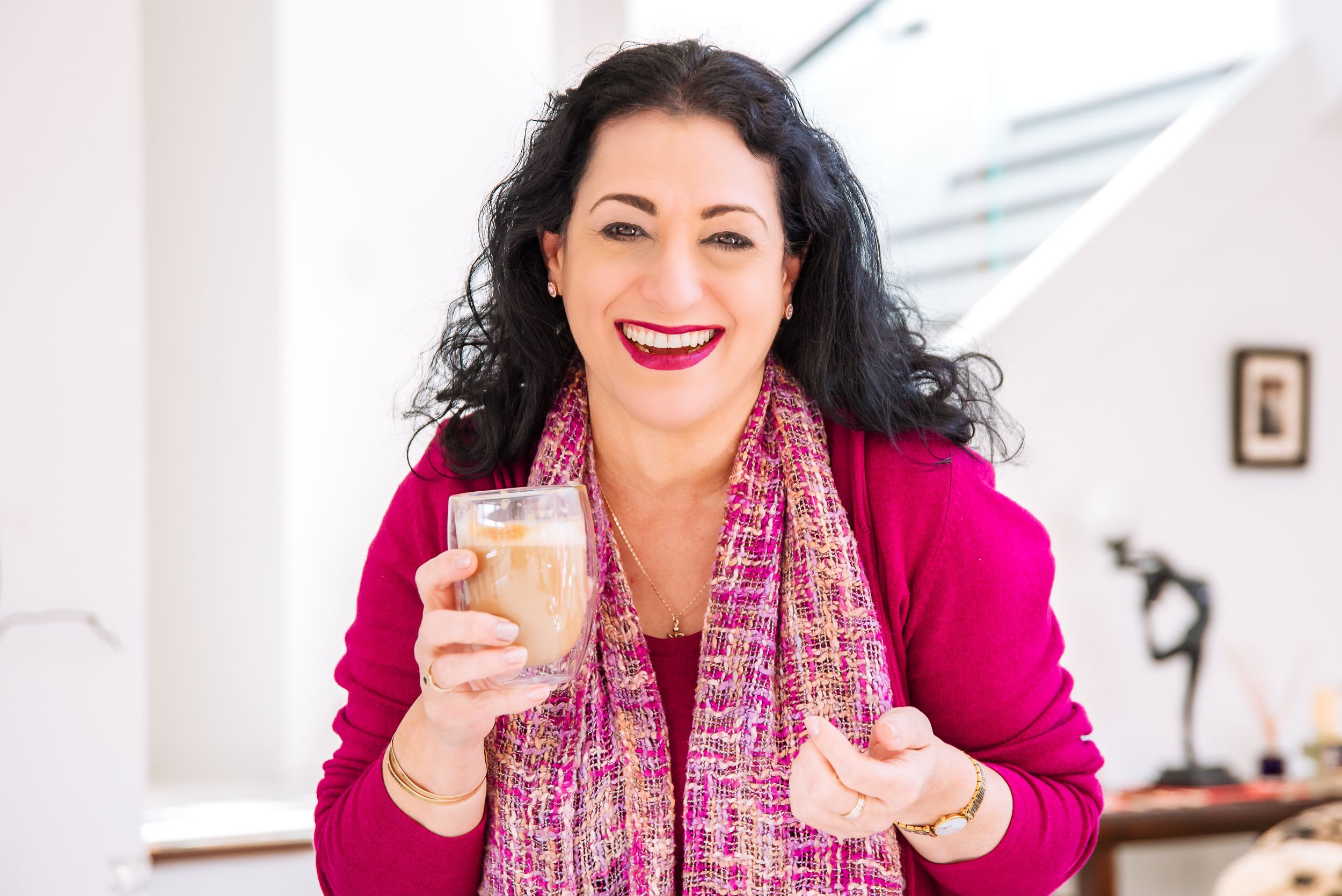 A person in a bright pink blazer smiles while holding a glass in a bright white room setting.
