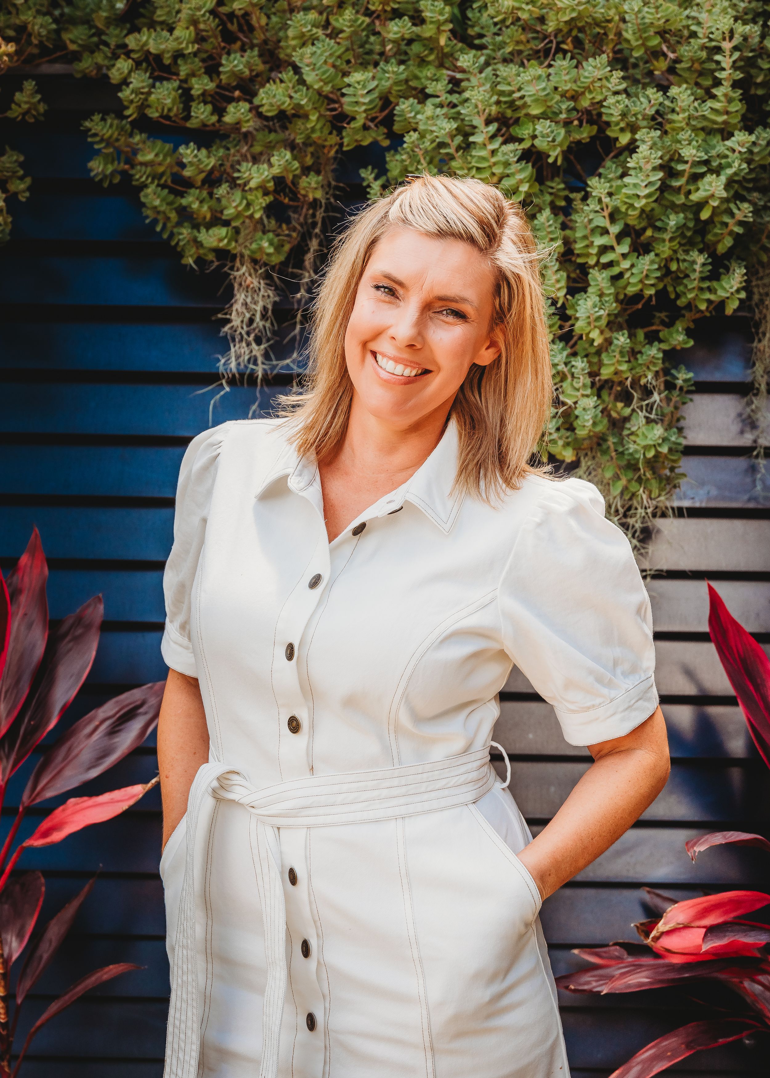 A person in a white button-down dress poses against a blue background with red-leafed plants visible.