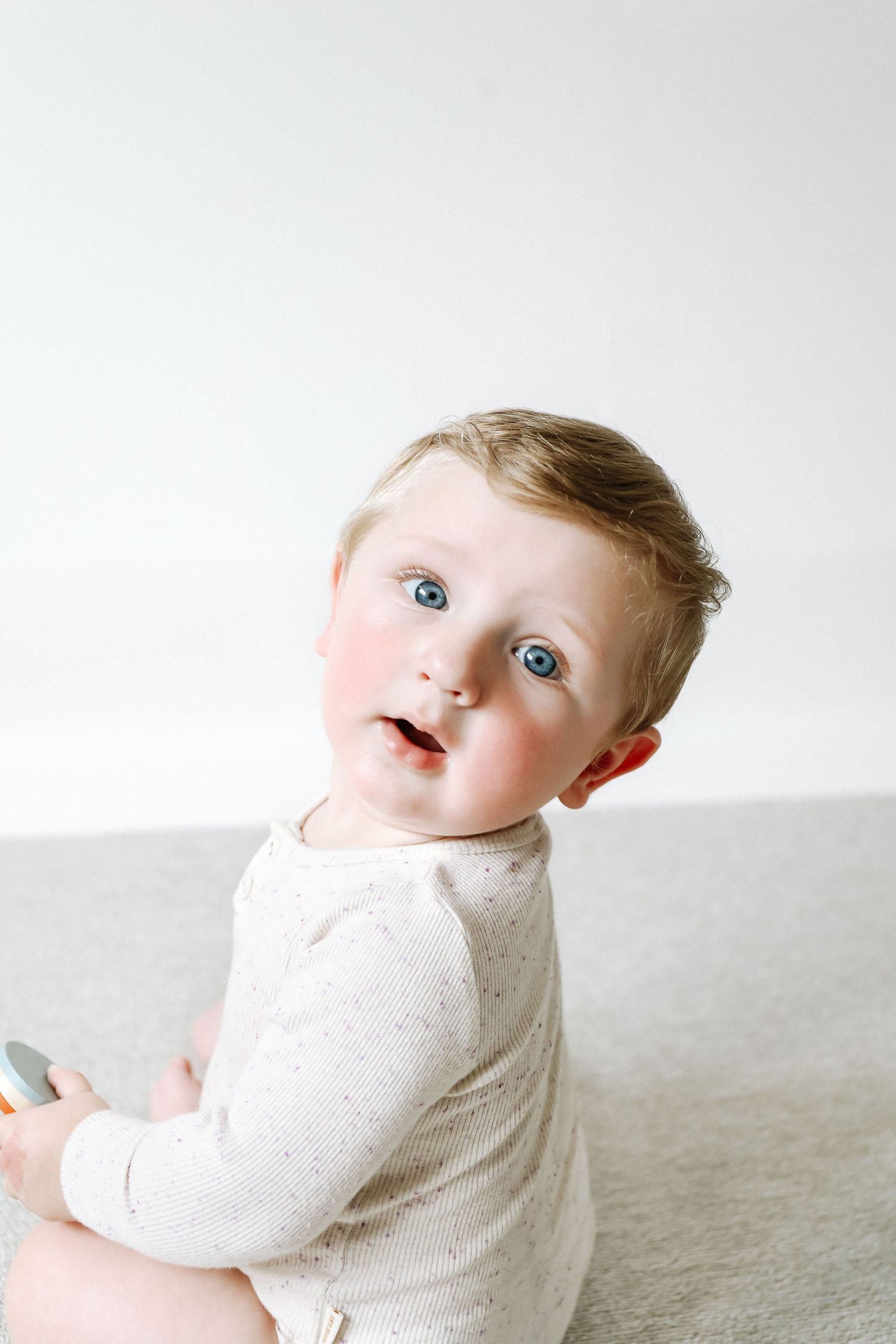 A baby in a white knit sweater looks back over their shoulder while sitting on a light grey surface during birthday shoot in Nottingham