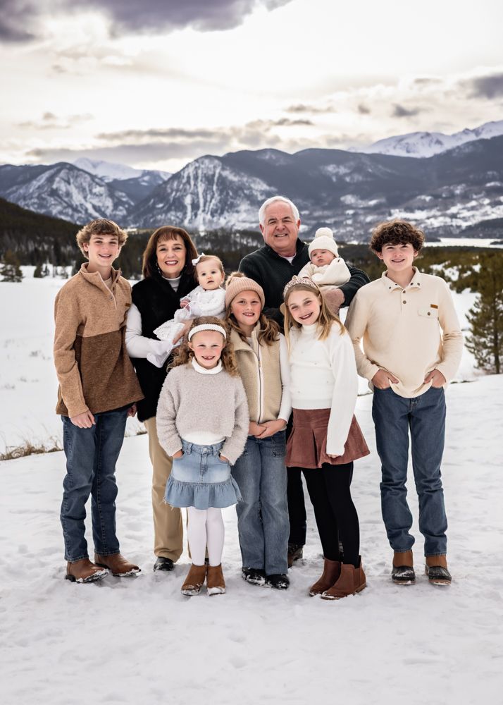 Multi-generational family of nine poses together in snowy mountain landscape during a winter outdoor photo session.