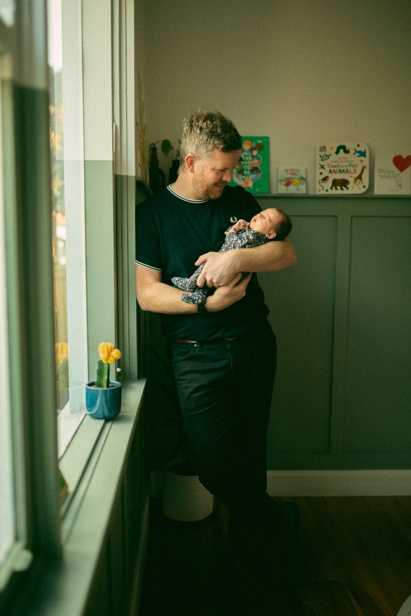 Someone holds a sleeping newborn baby near a window with a yellow tulip on the windowsill and green wainscoting on the wall.