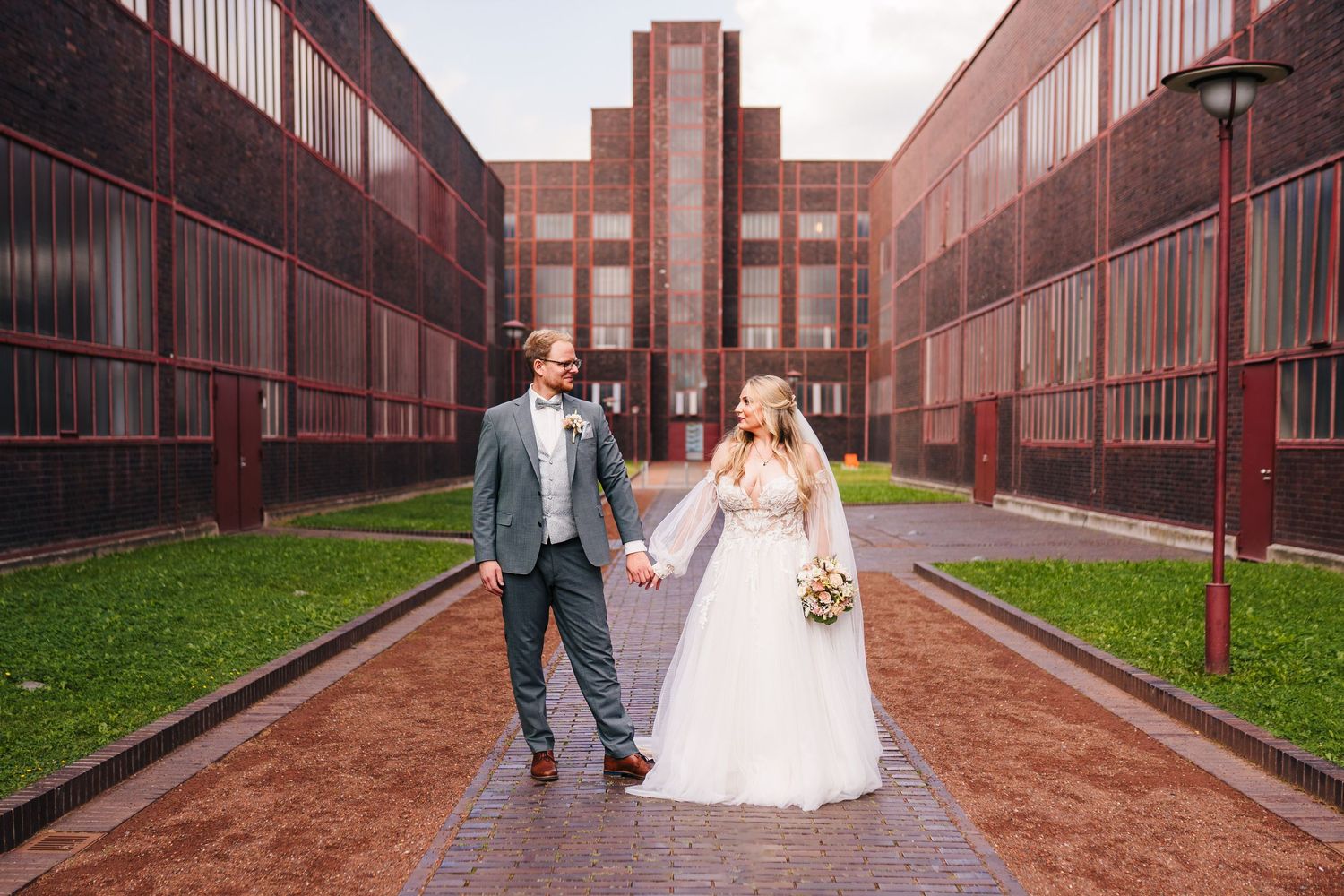 A couple walks hand in hand down a brick pathway between red buildings on their wedding day.