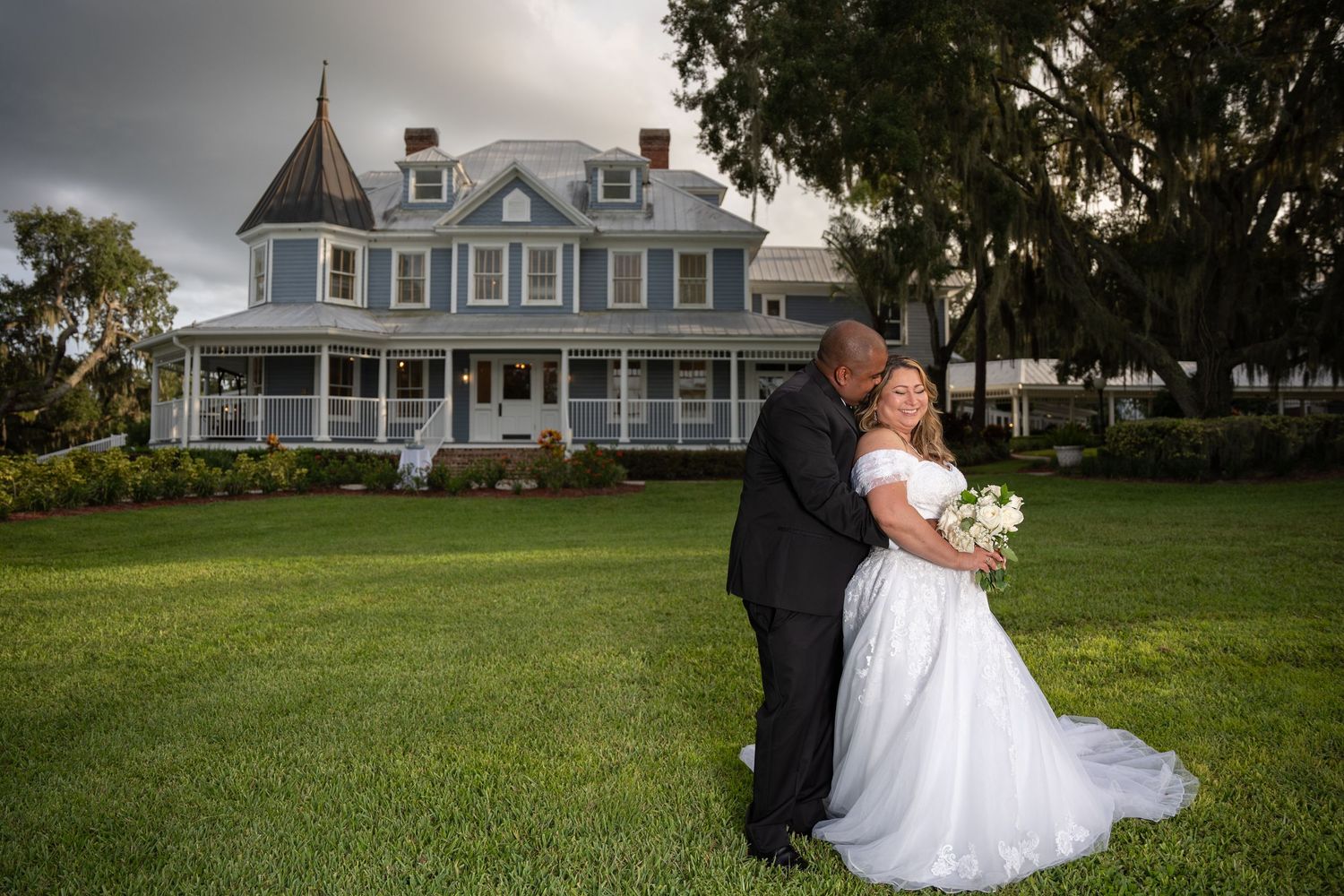 Bride and groom share tender kiss on lawn before charming Victorian house after wedding ceremony.