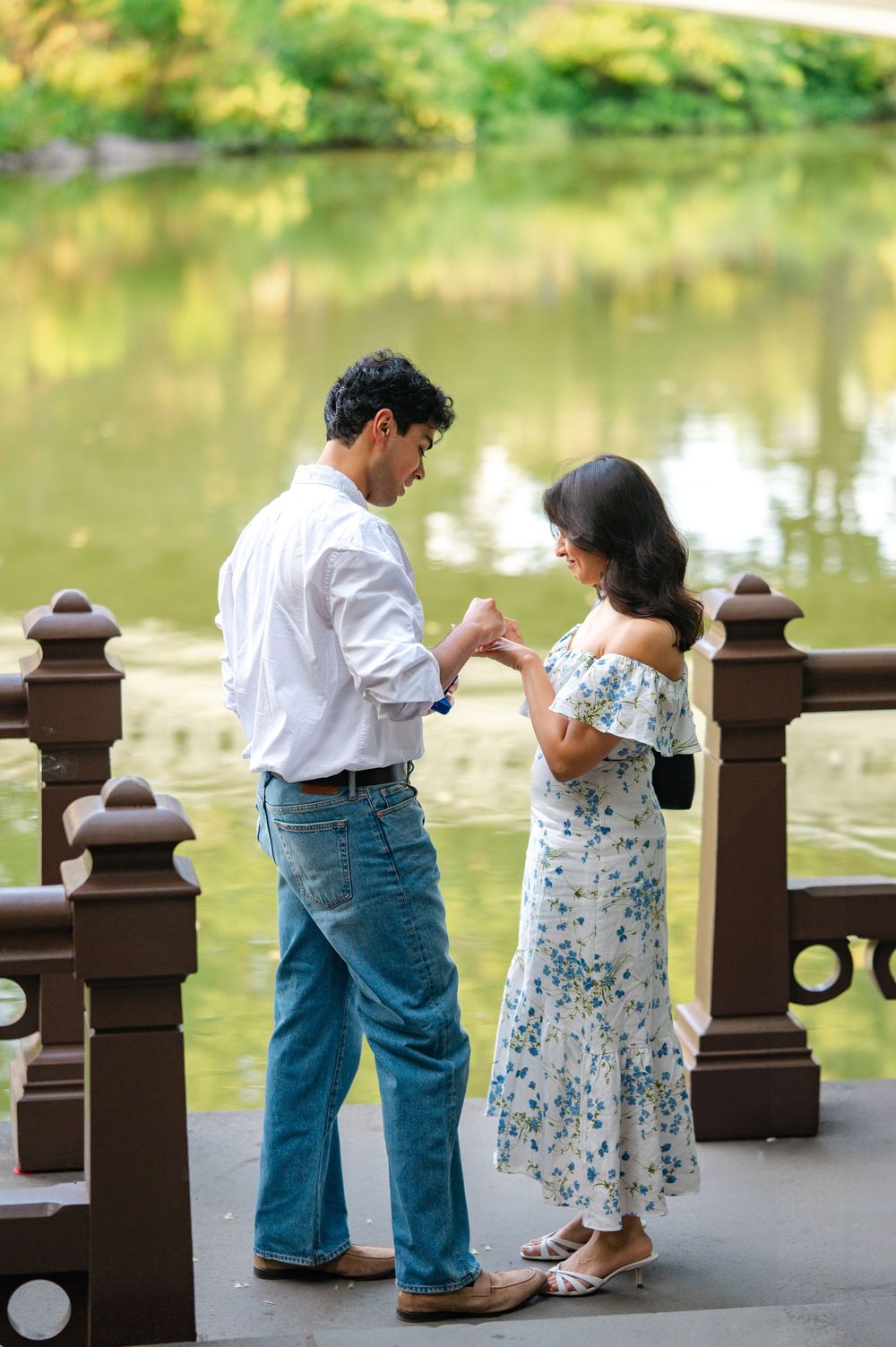 A man slides an engagement ring on a woman's hand on a bridge overlooking the serene Central Park Lake with green reflections in the water.