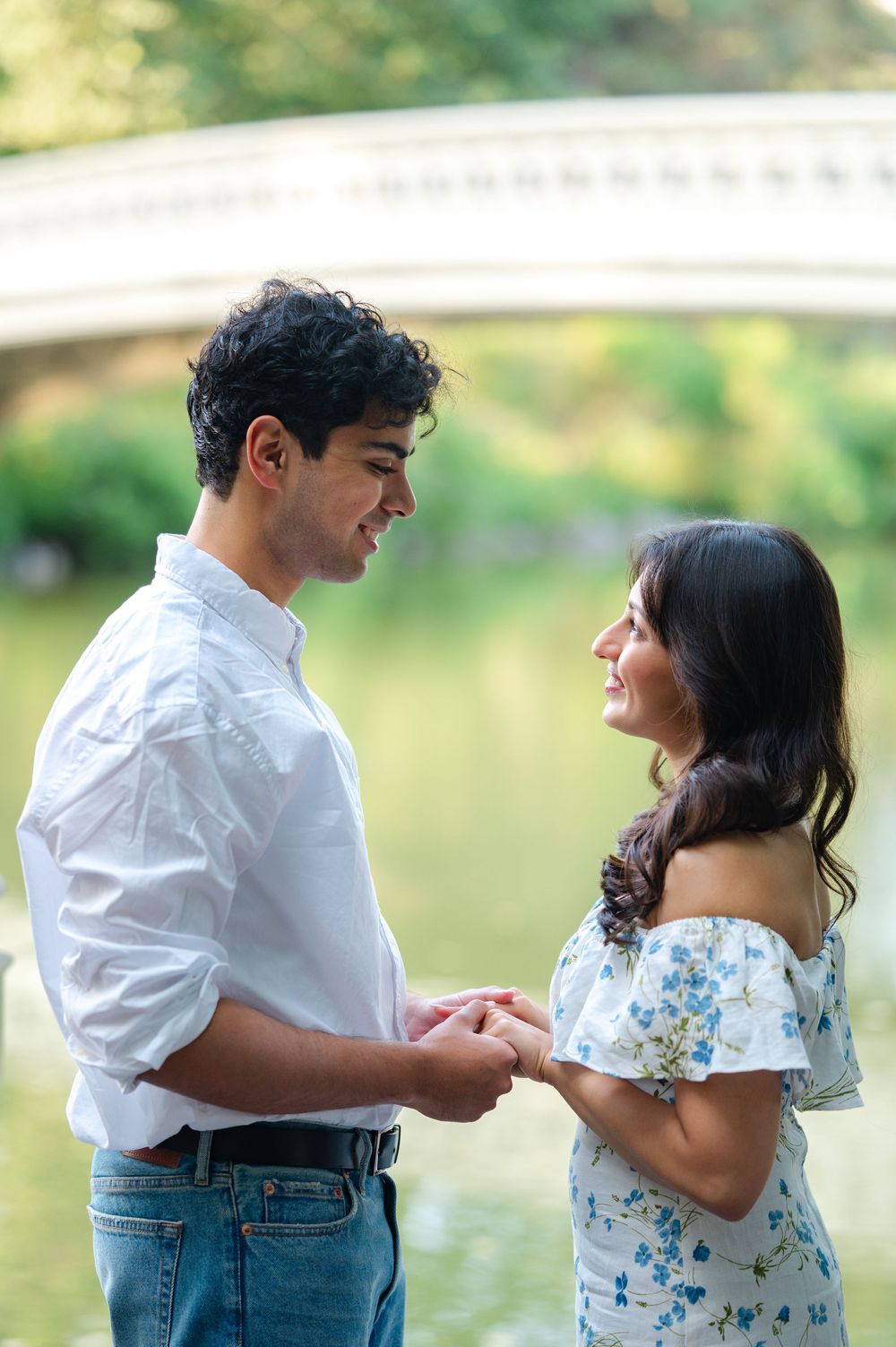 A couple in white and floral clothing holds hands by Central Park Lake in a series of sweet engagement photos after their proposal