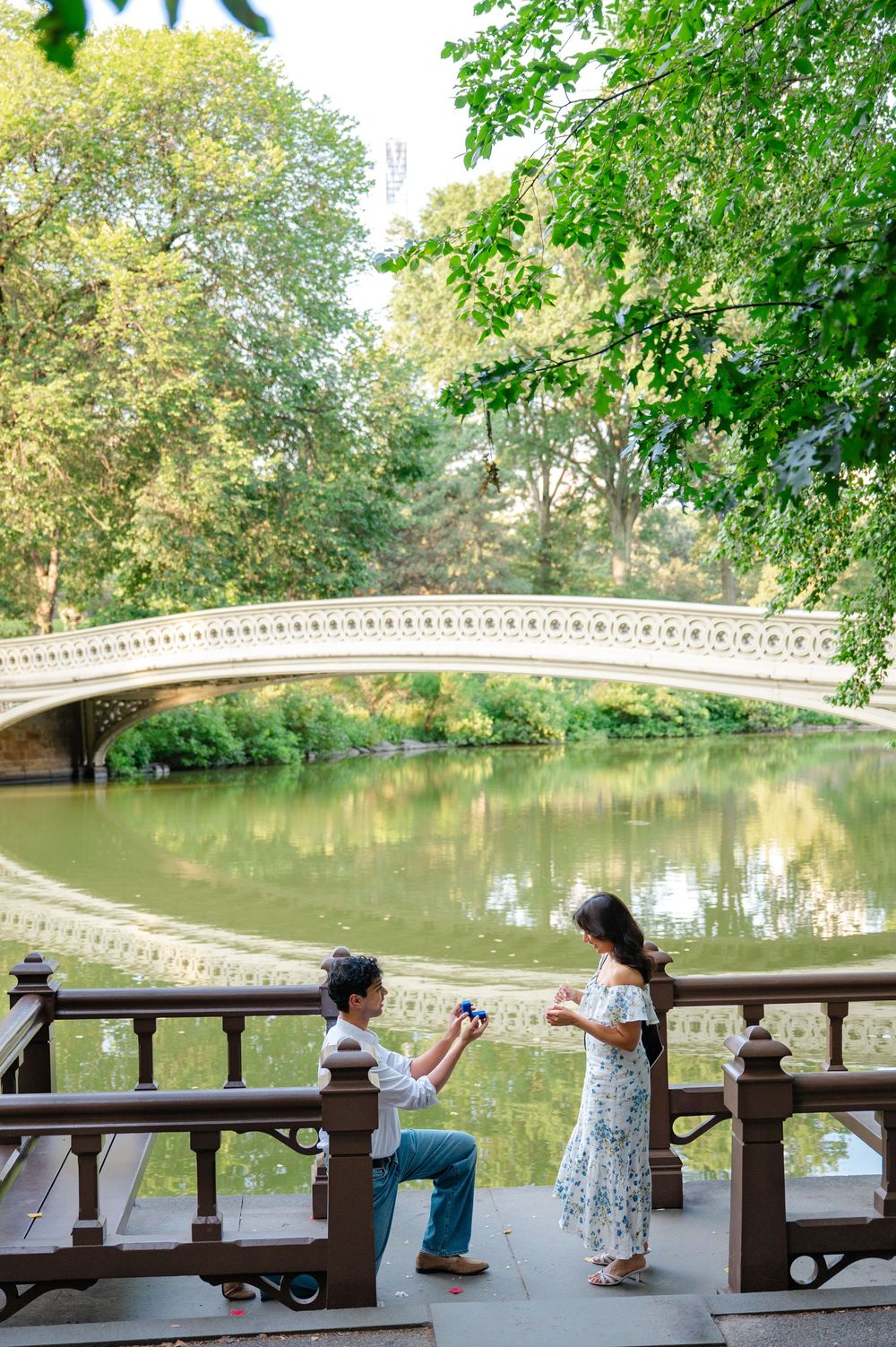 A romantic proposal moment unfolds near Bow Bridge in Central Park with the iconic arch bridge reflecting in the lake.