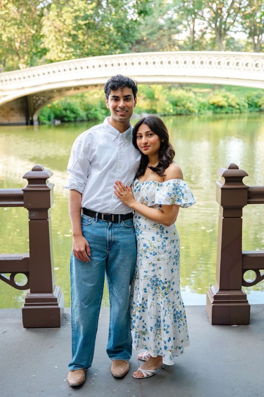 A couple poses together in front of Bow Bridge in Central Park, wearing casual summer attire against a scenic backdrop after their proposal