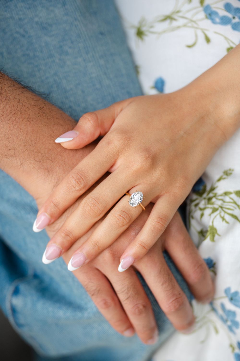 A delicate engagement ring with a solitaire diamond sparkles on a hand against a backdrop of floral patterned fabric.