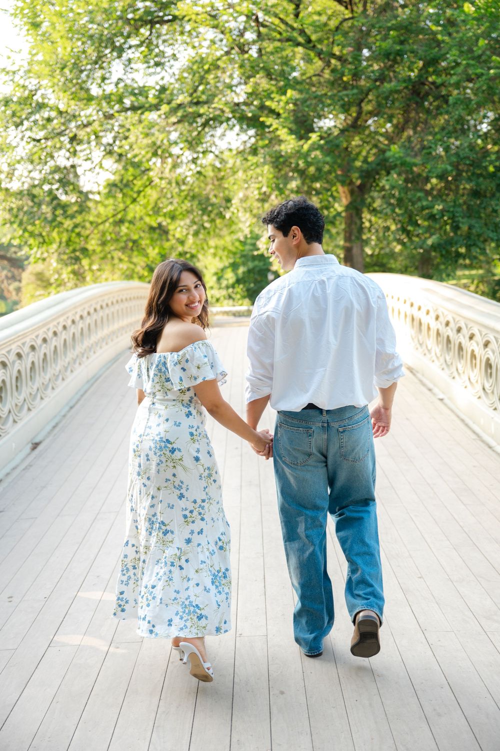 A couple walks across the Bow Bridge in Central Park together after getting engaged. The woman is looking back while the man looks at her.