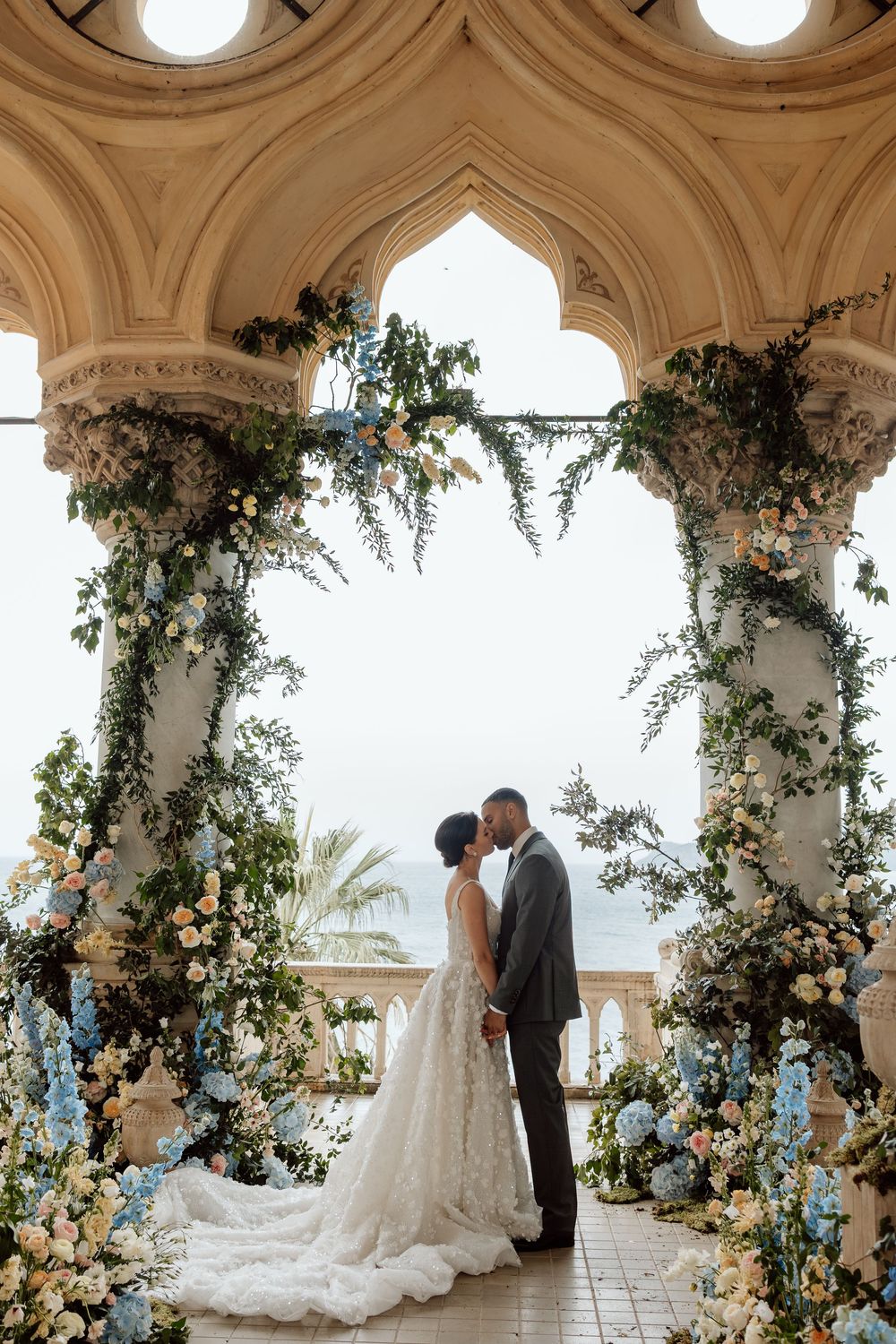 Romantic wedding ceremony under ornate stone archway decorated with blue and white floral arrangements and greenery.