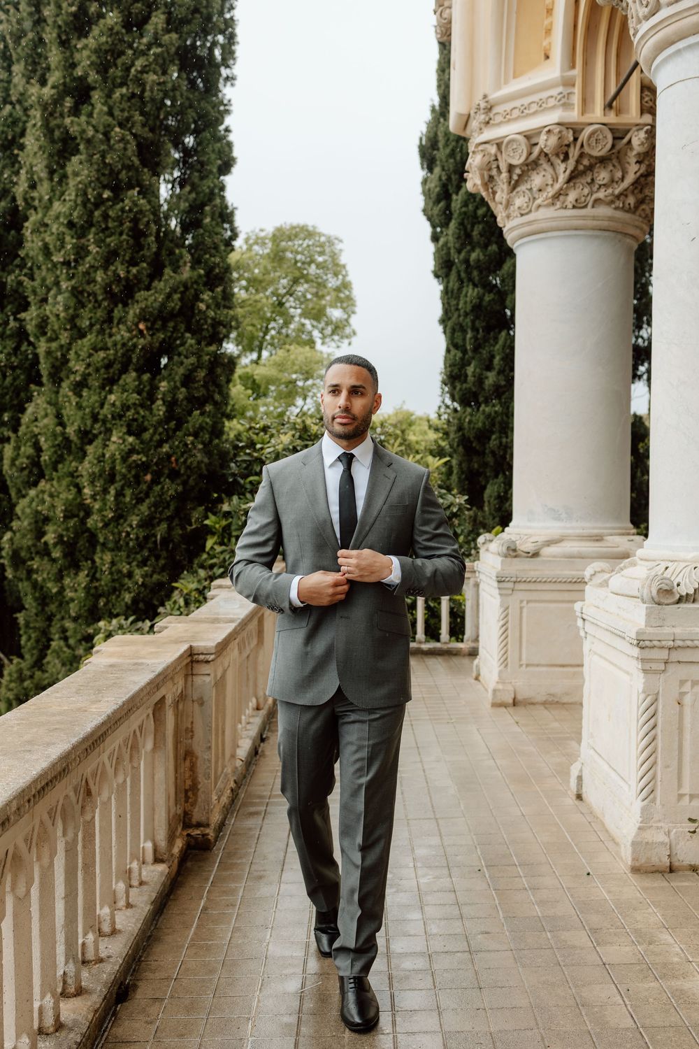 Professional in a grey suit poses elegantly against ornate white columns and balustrade in an outdoor setting.