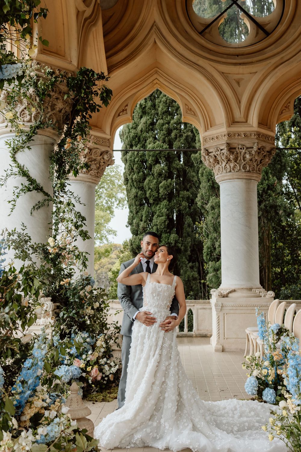 A couple embraces under ornate stone archways decorated with white and blue floral arrangements at an elegant venue.