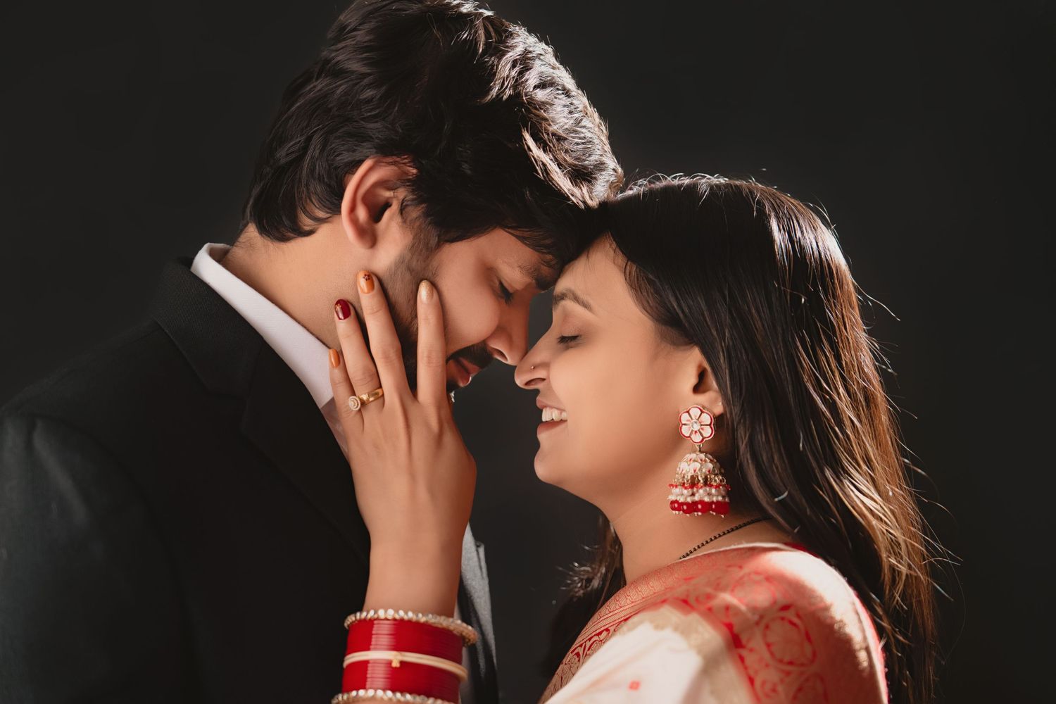 Romantic couple in traditional South Asian attire share an intimate moment as she wears a red sari and jewelry.