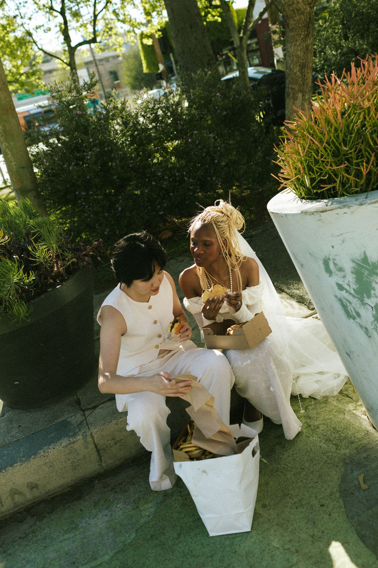 A candid shot of two brides sit on a curb in their suit and dress, snacking on some cheeseburgers and fries