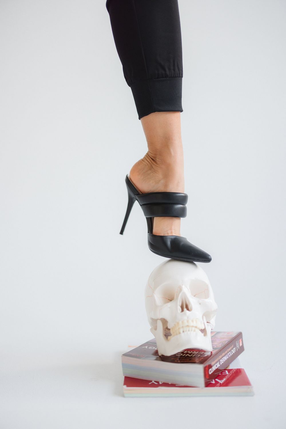A stiletto heel balanced on a white skull and stack of books in a minimalist fashion photograph against light background.