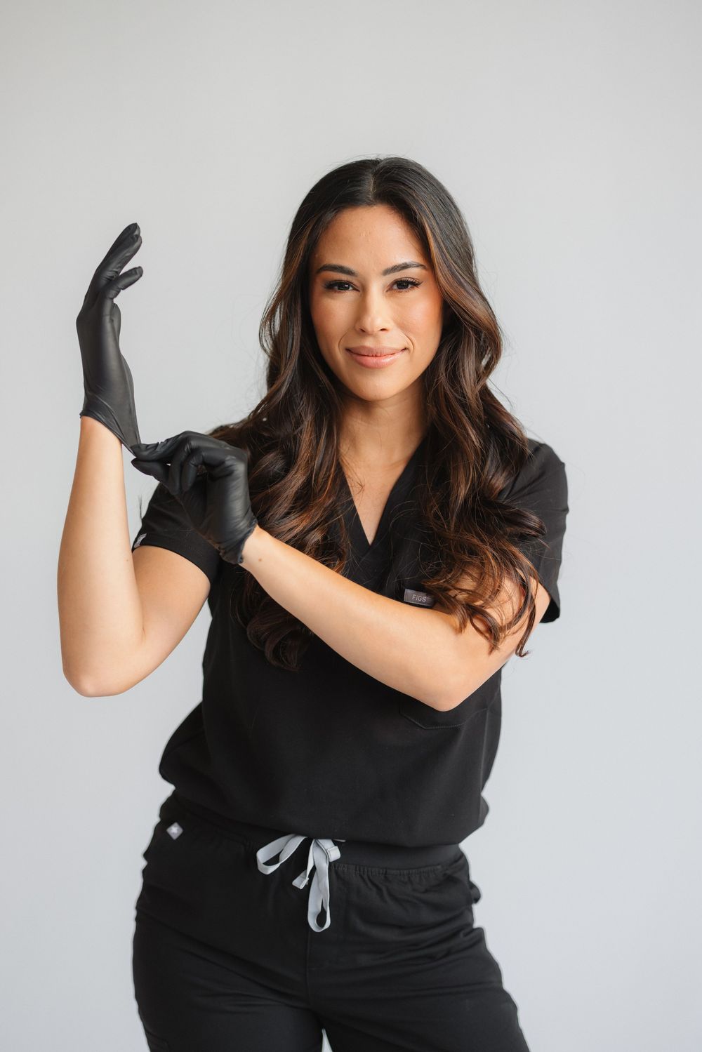 Healthcare professional in black scrubs and gloves poses confidently against light background during portrait session.