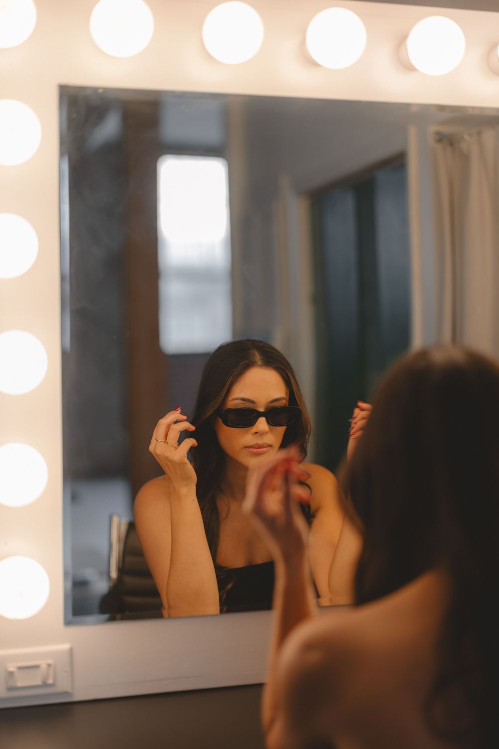 Woman wearing black sunglasses looking in illuminated vanity mirror with round bulbs in dressing room.