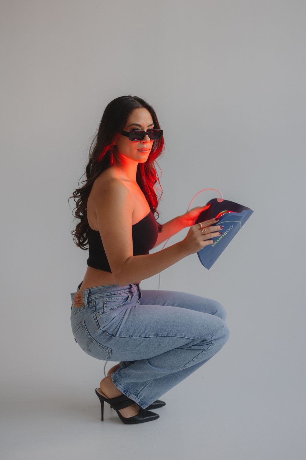 Woman in black crop top and light jeans crouches down while holding book with red lighting effect in studio setting.