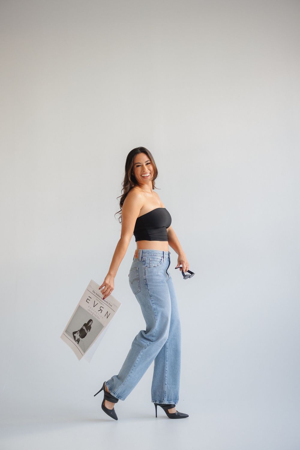 A woman in high-waisted jeans and black top poses with a shopping bag against a white backdrop in a dynamic series.