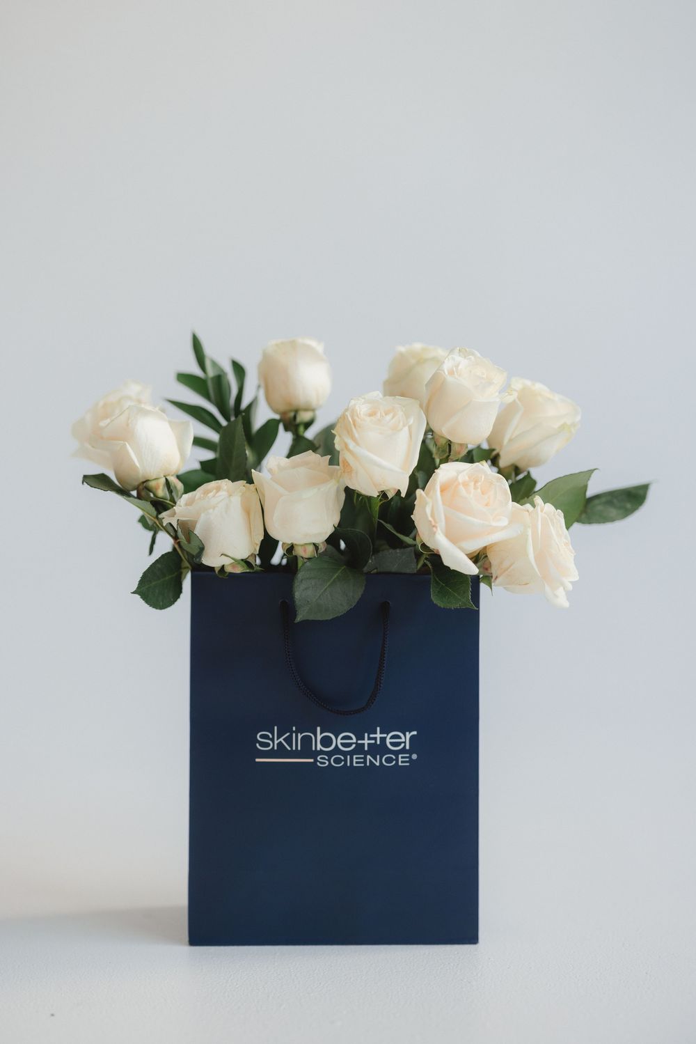 White roses arranged in a navy blue Skinbetter Science shopping bag against a light gray background.