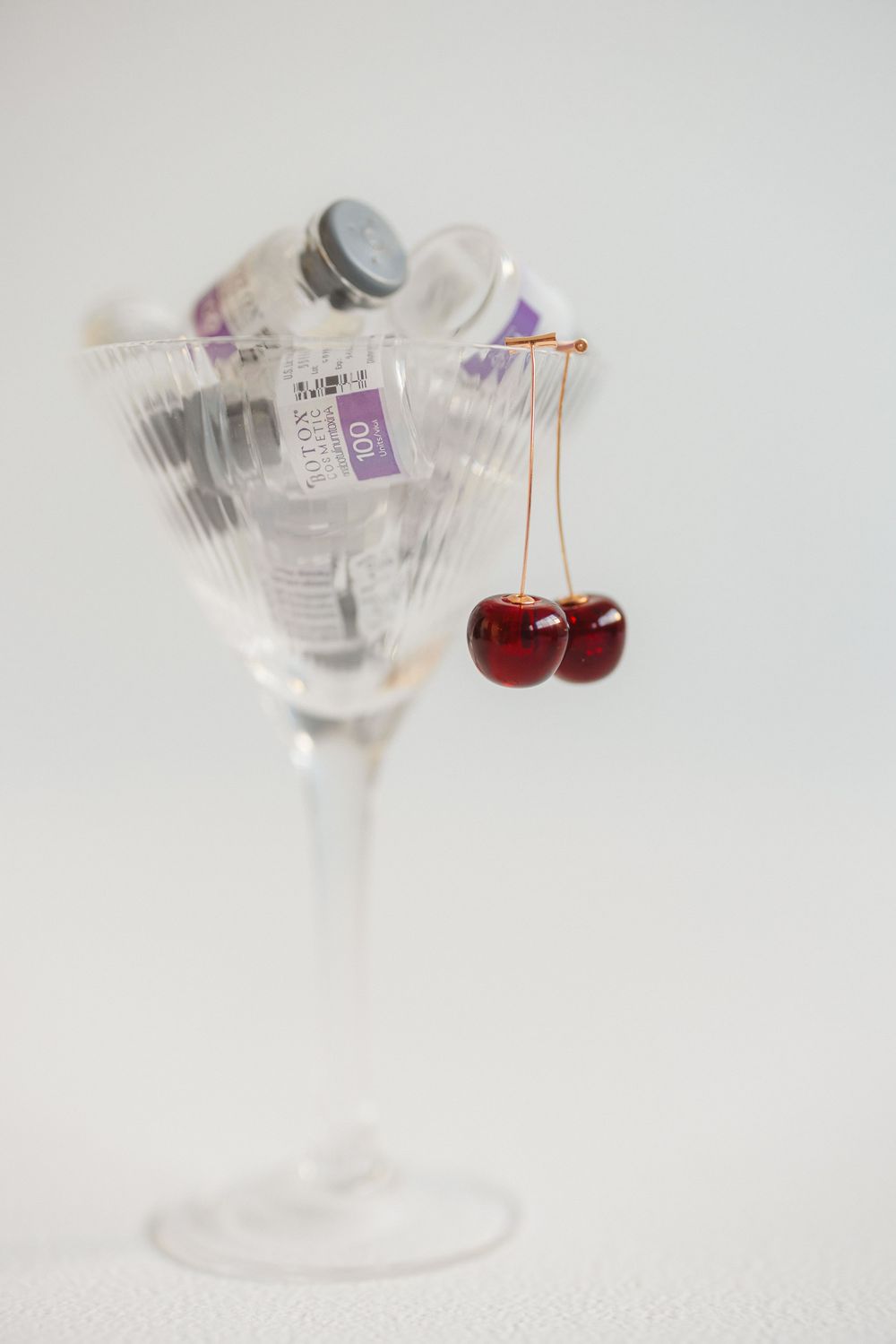 A clear glass martini glass with two red cherries hanging on the rim against a white background.