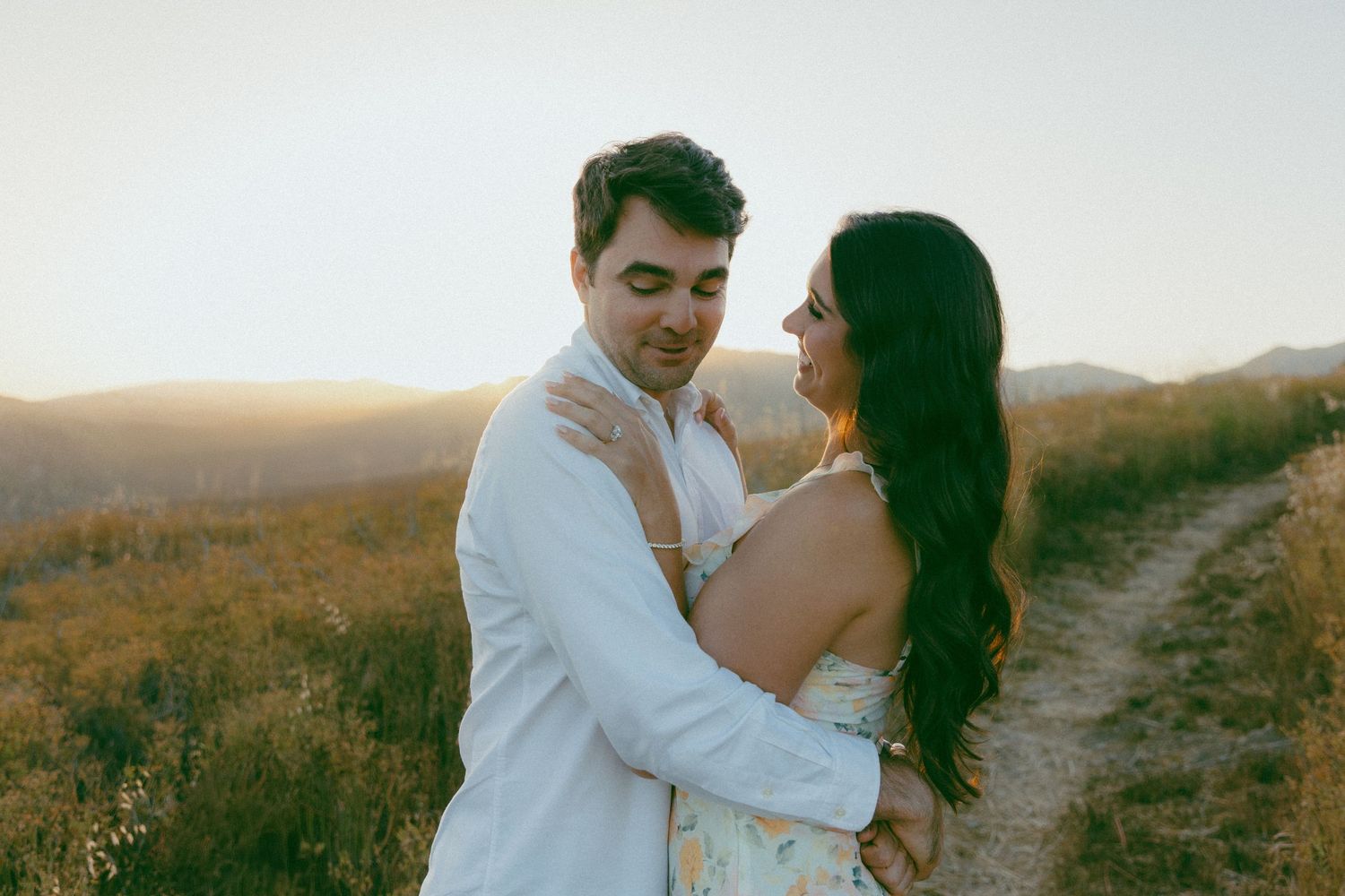 A romantic couple embraces on a hilltop trail during golden hour with scenic rolling hills in the background.