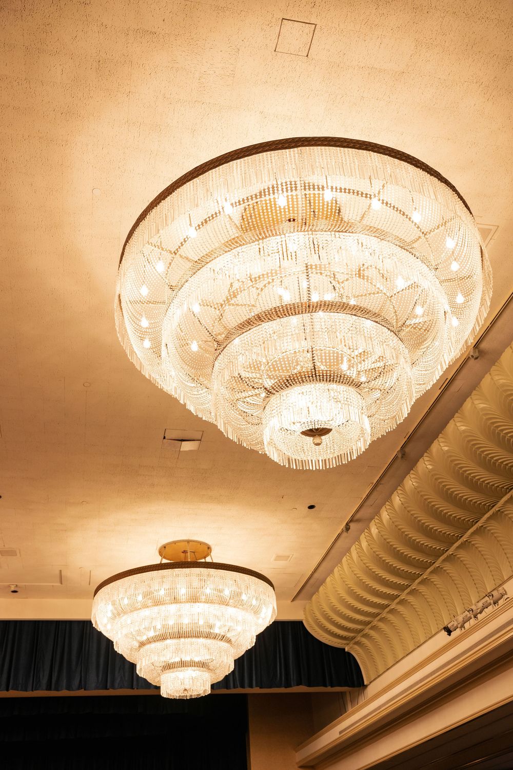 Elegant tiered crystal chandeliers with warm lighting illuminate an ornate ballroom ceiling with exposed ductwork.