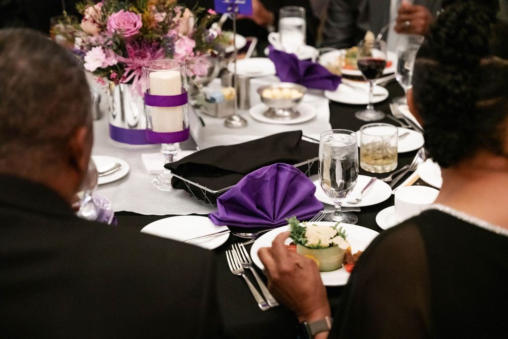 An elegant wedding reception table setting with purple napkins, white plates, and pink floral centerpieces.