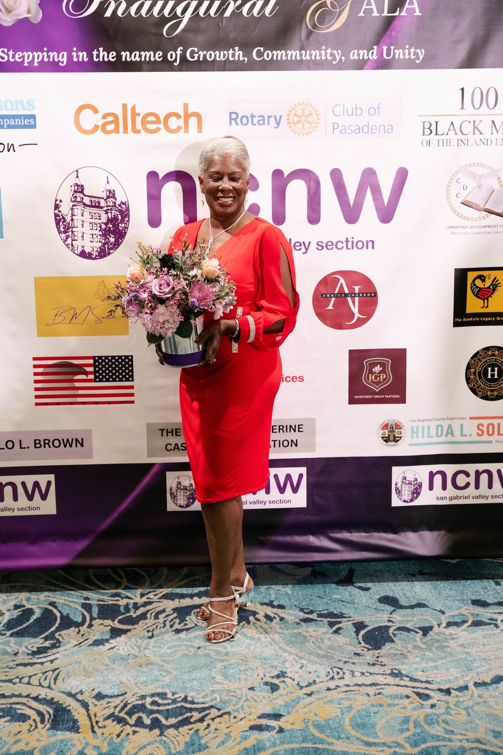 A beaming person in a vibrant red dress holds a beautiful purple floral arrangement while standing in front of a sponsor wall.