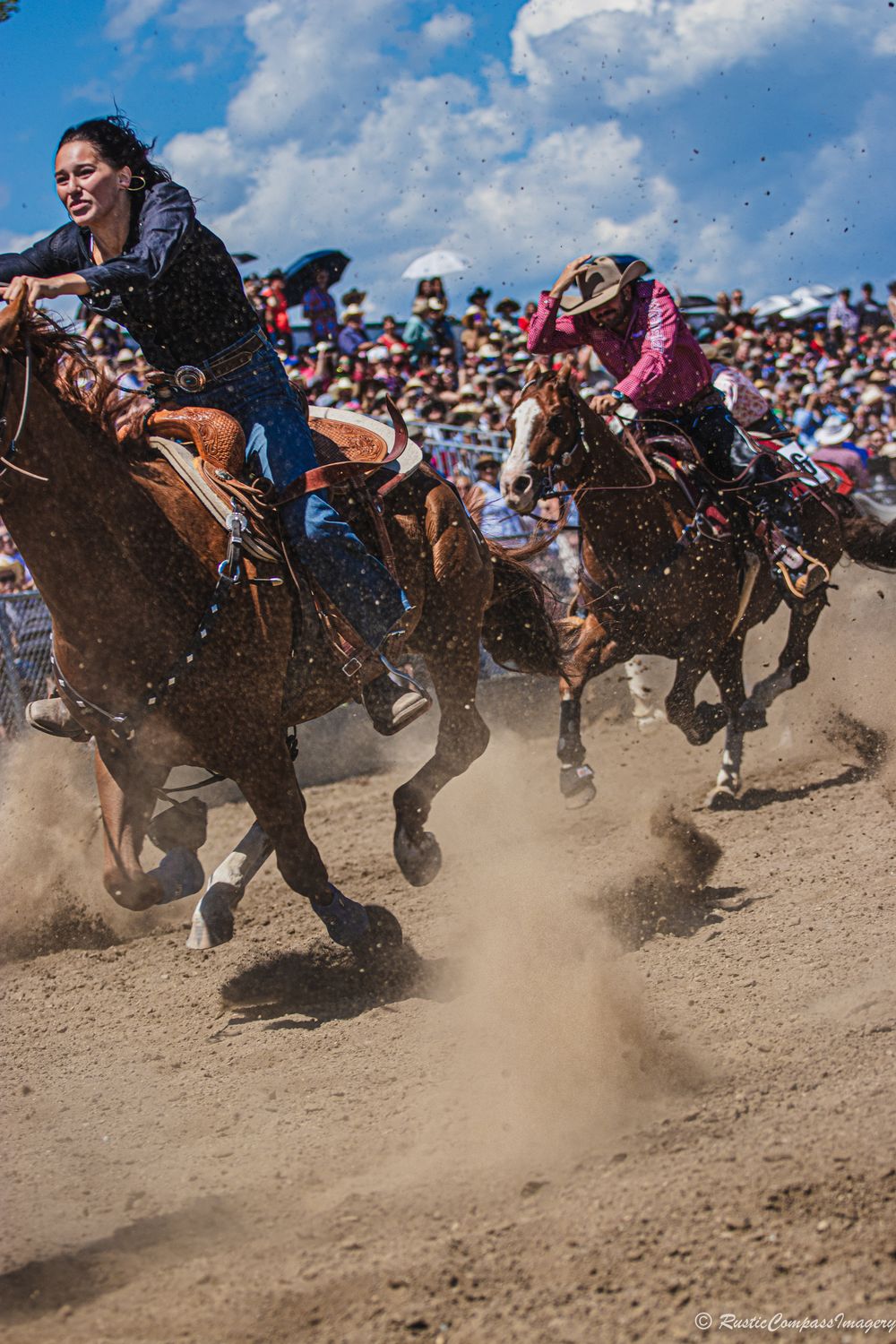 Multiple horse riders compete in a fast-paced racing event kicking up dust on a dirt arena track under blue skies.