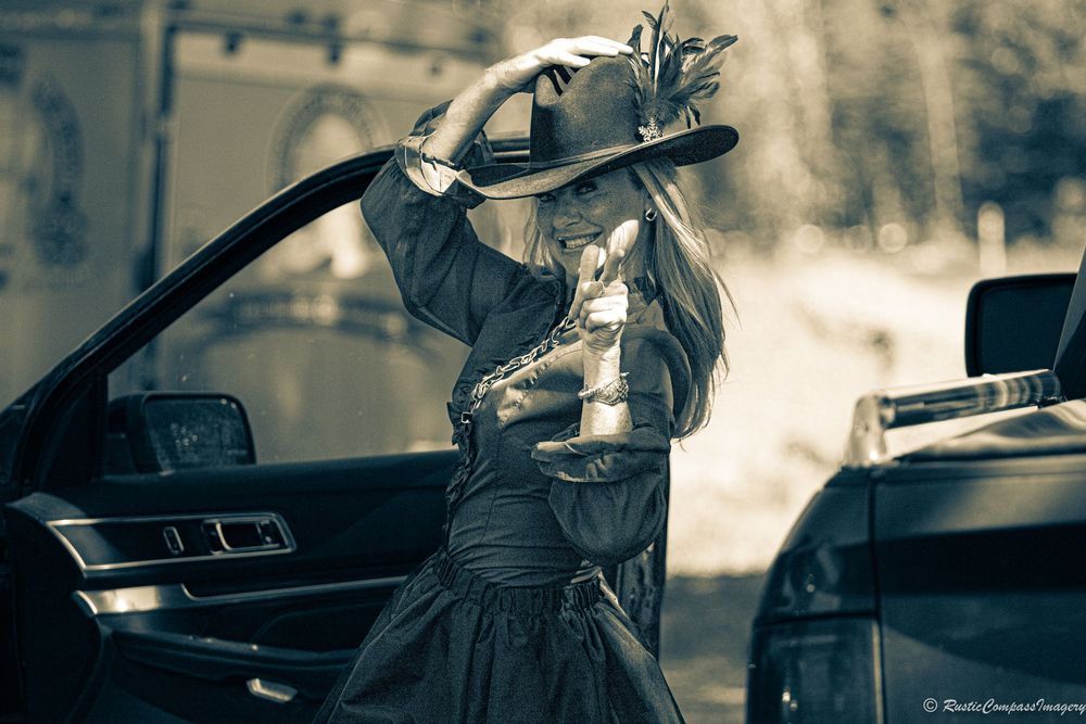 A person in side saddle attire with a top hat stands by an opened car door in monochromatic tones.