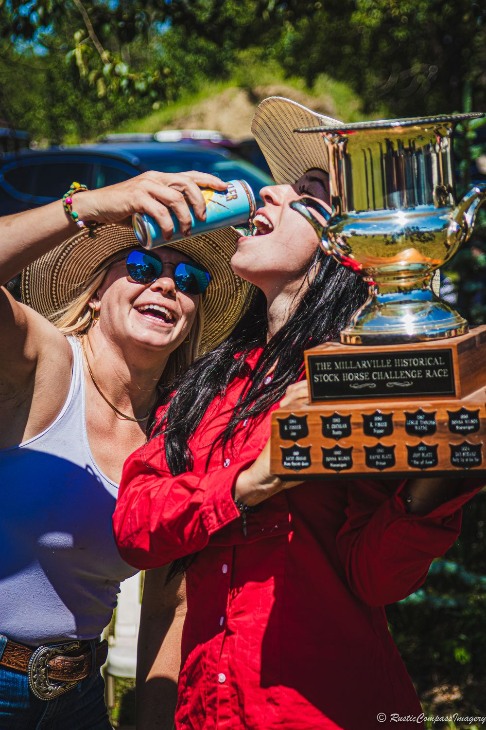 Two jubilant people celebrate with a large trophy and drinks at an outdoor summer event.