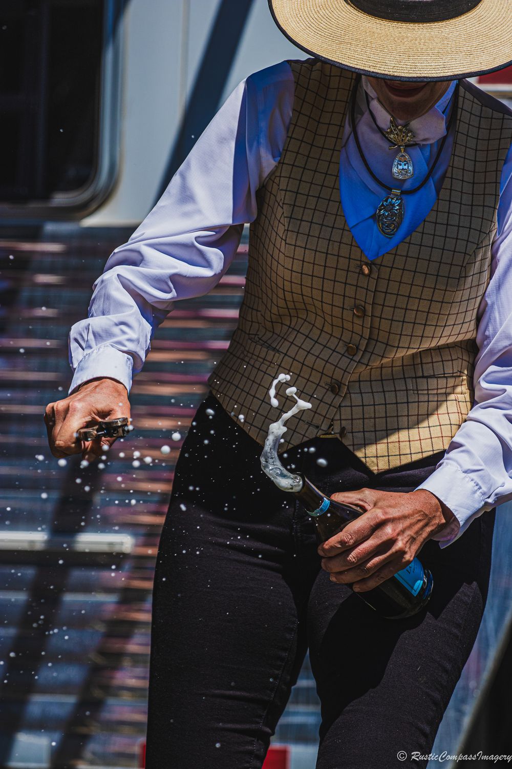 Person wearing western attire with checkered vest and straw hat opens a bottle of champagne using a knife