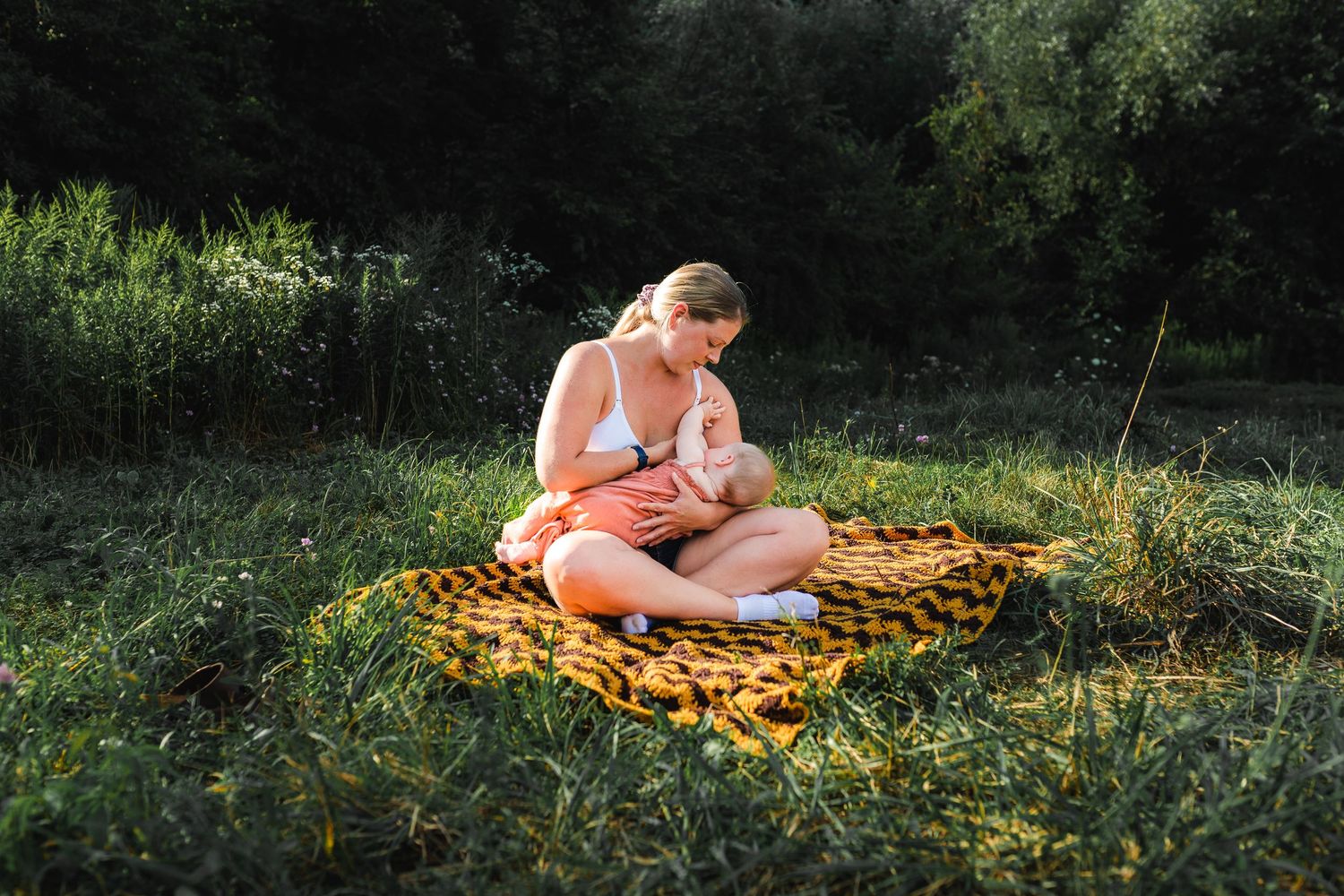 Mother sits on an orange blanket in a grassy meadow on a sunny summer day, breastfeeding infant.