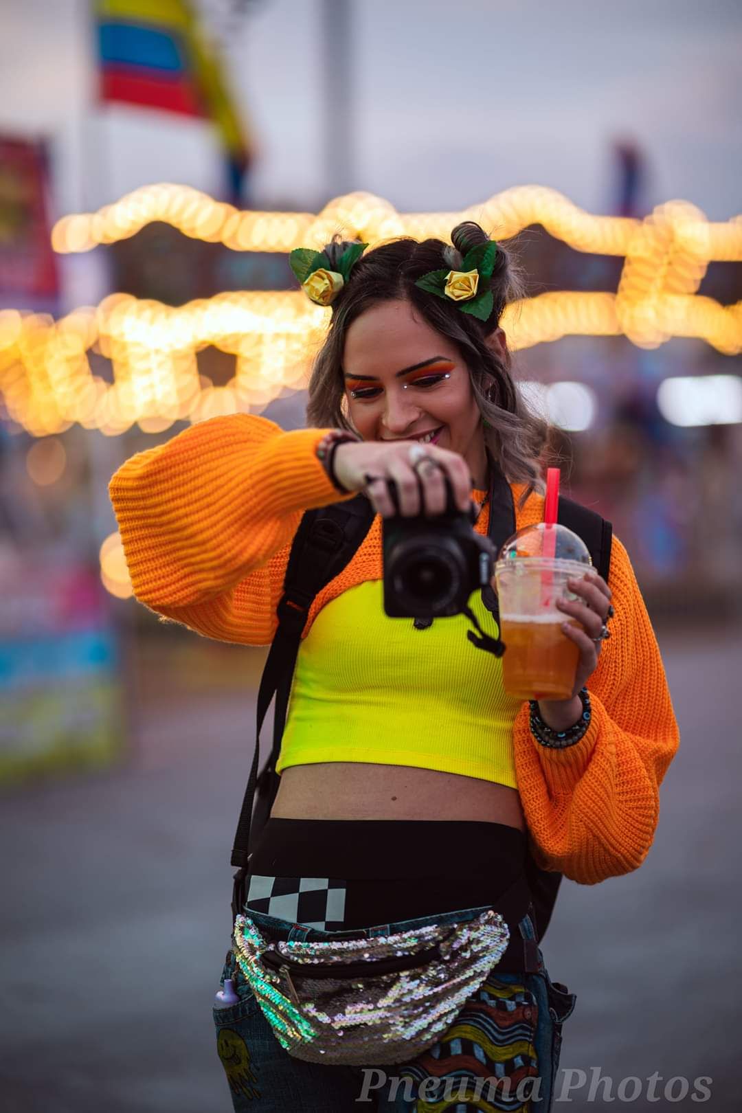 Smiling woman in orange sweater and neon yellow crop top holding camera and drink at a colorful festival.