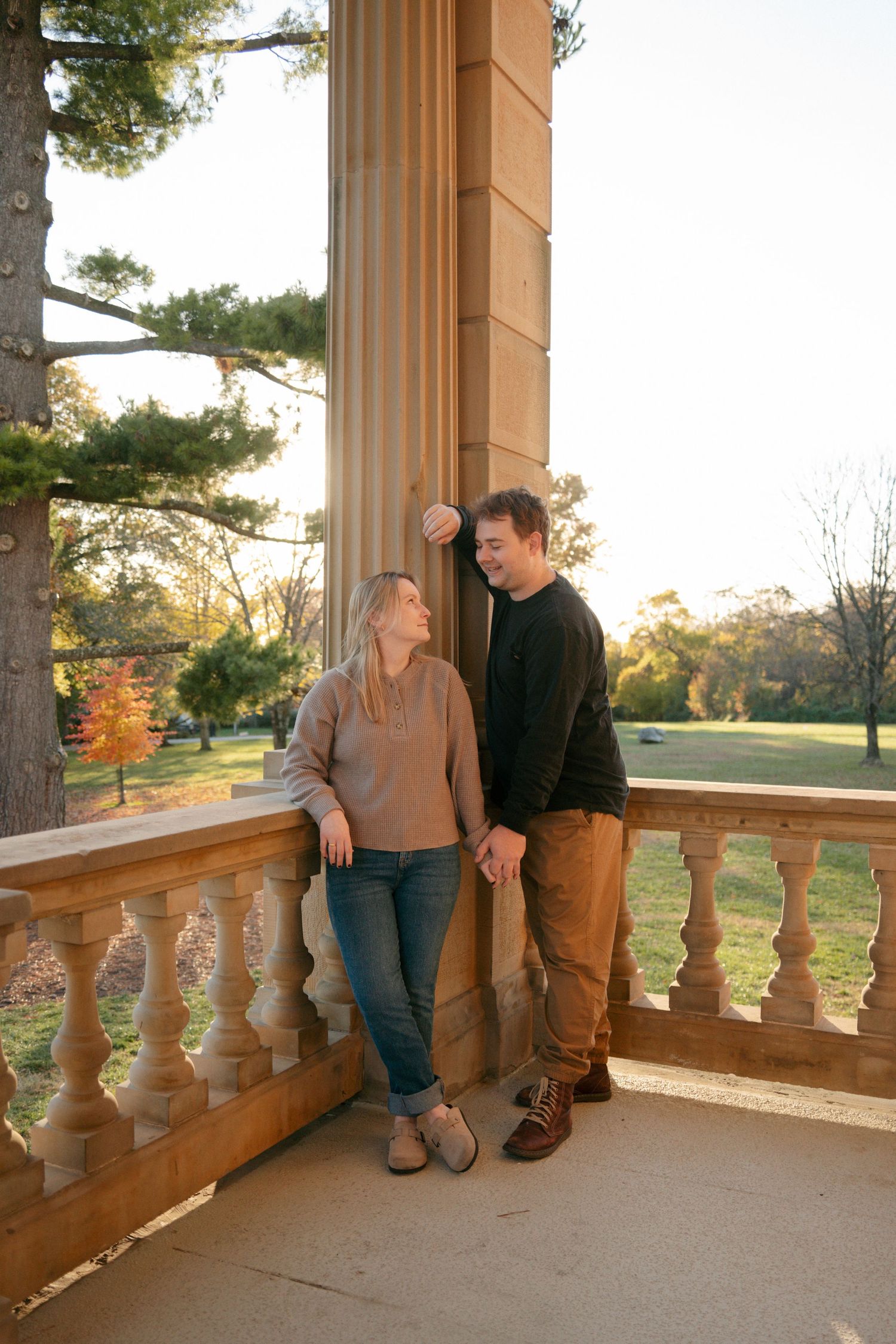 Couple holding hands and smiling on sunlit stone balcony surrounded by autumn park scenery.