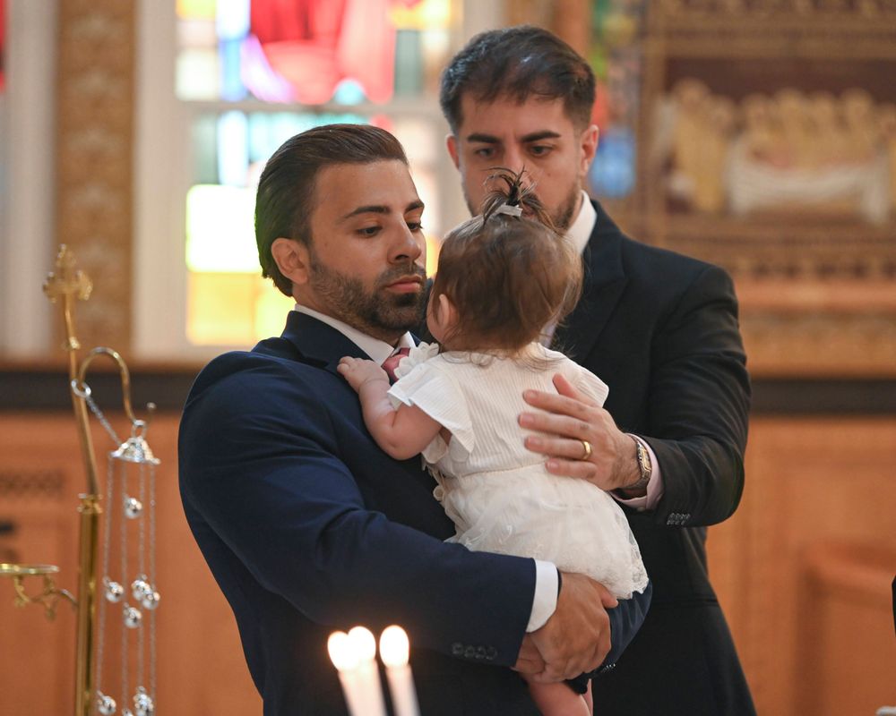 Baby in white christening gown being held during religious ceremony with candles.