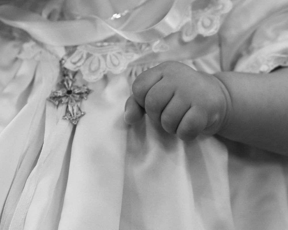 A small baby's hand gently rests on delicate white lace fabric in a black and white photograph.
