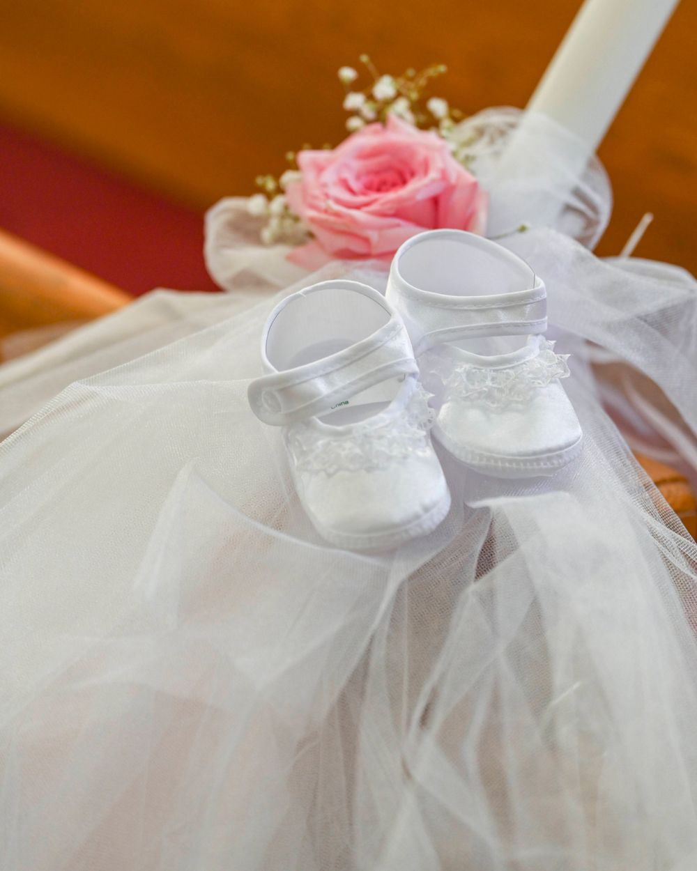 Delicate white baby shoes with lace details displayed on tulle fabric next to a pink rose.