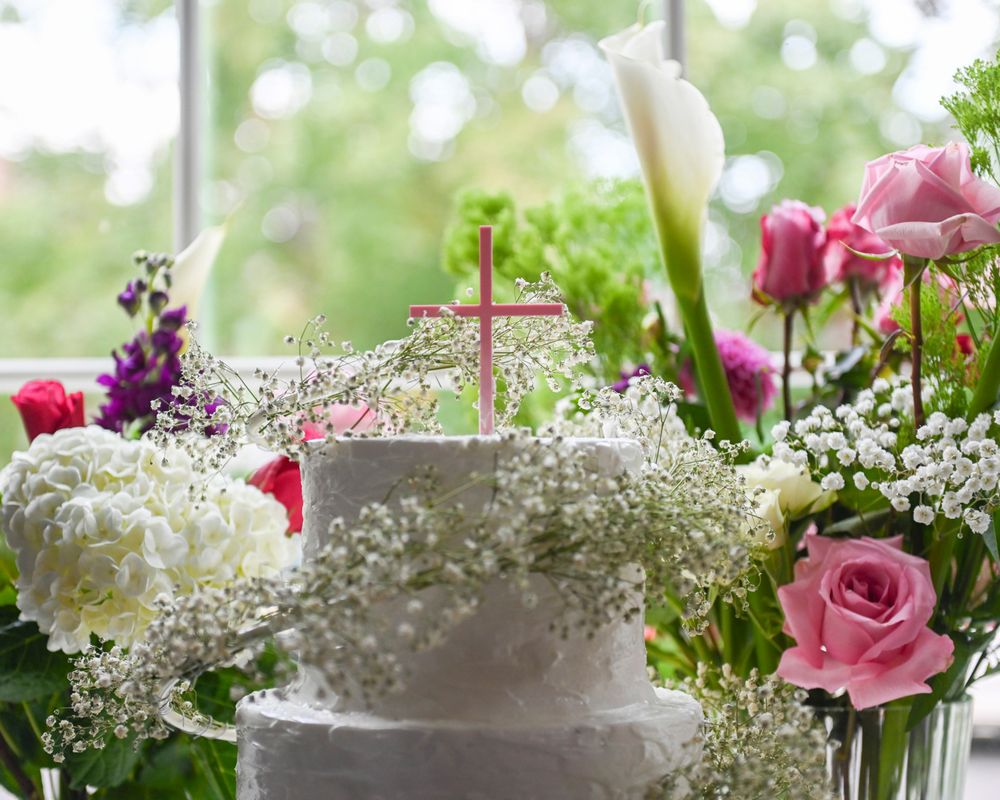 An elegant christening cake adorned with white flowers, pink roses, and baby's breath in a garden setting.