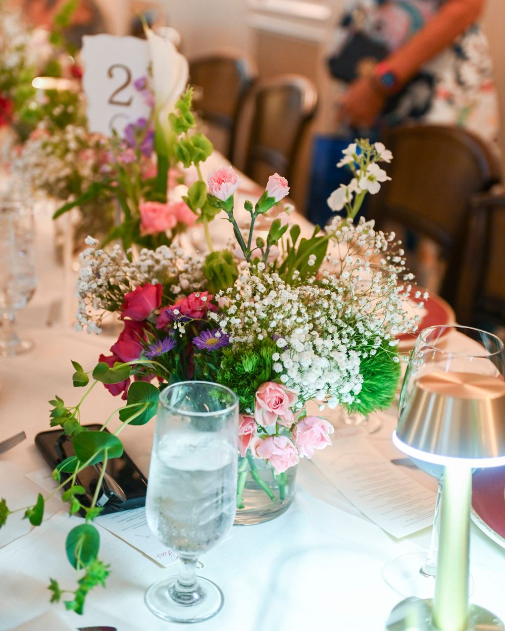 Table centerpiece with pink roses, baby's breath, and a table number card for a formal event.