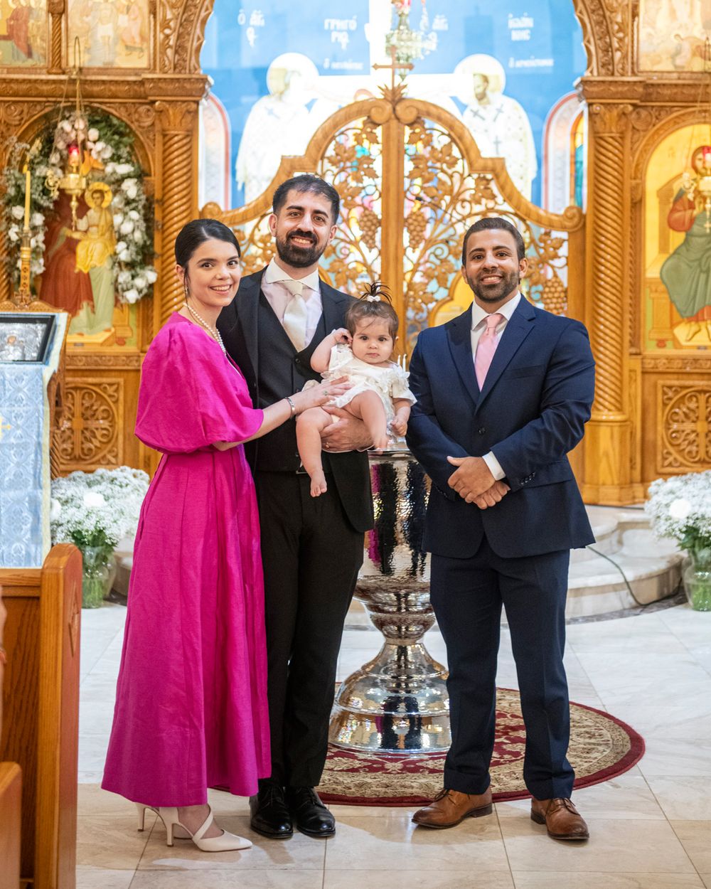 A baptismal ceremony taking place inside an ornate Orthodox church with decorative iconography and floral arrangements.