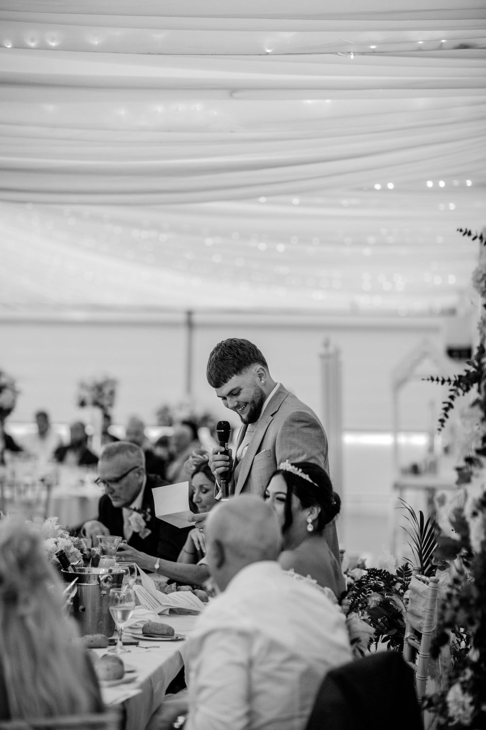 Black and white photo of a wedding reception speech under an elegant draped tent ceiling.