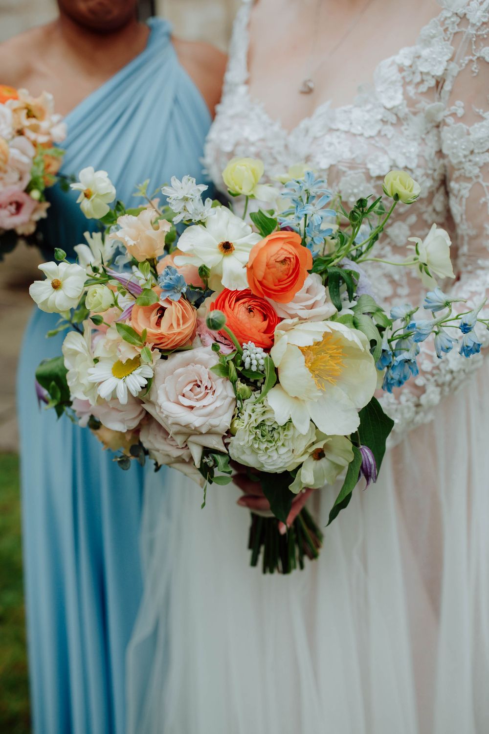 A wedding bouquet featuring orange ranunculus, white daisies, and blue delphinium against light blue bridesmaid dress. Blackfriars Priory