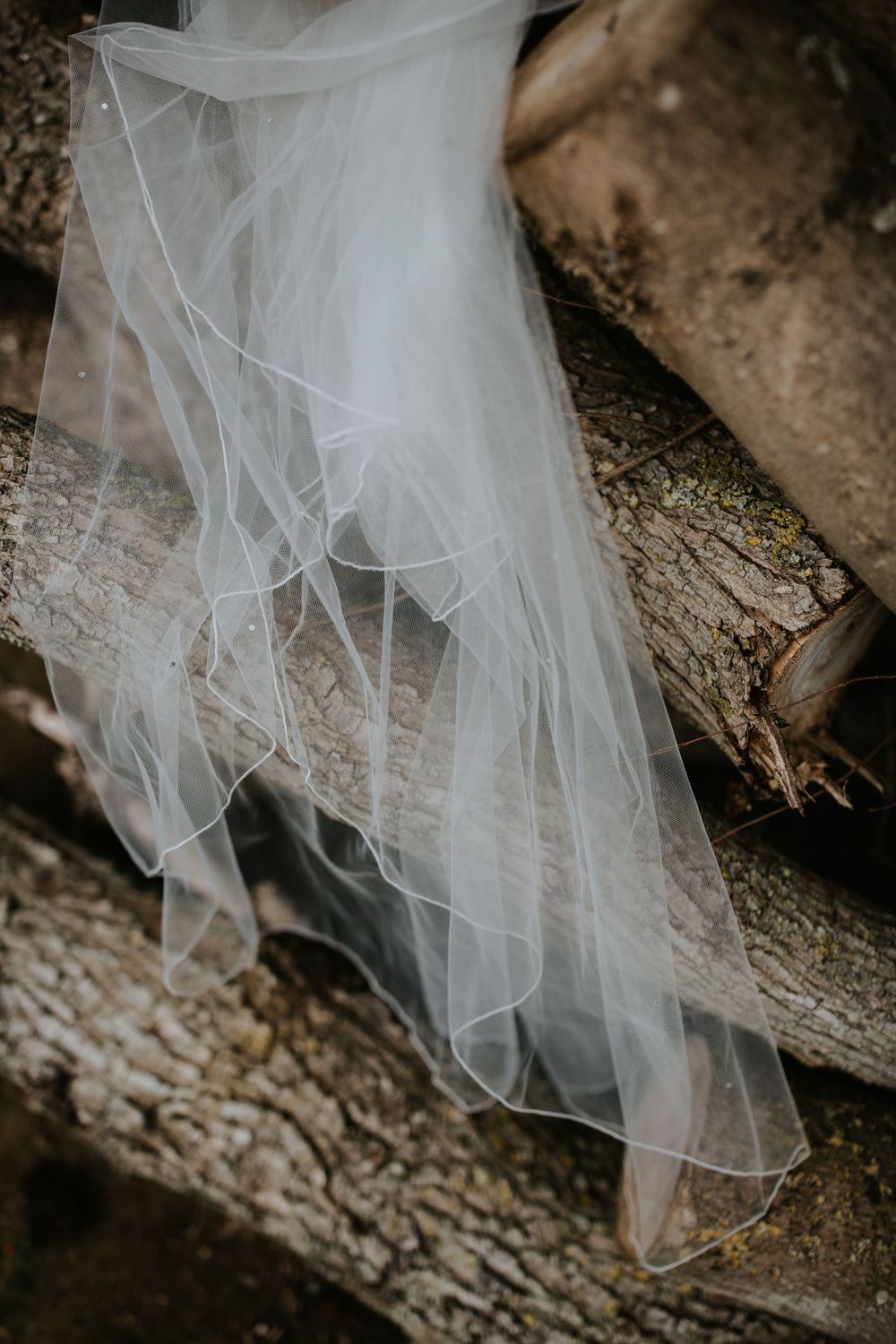 Delicate white wedding veil draped over rustic wooden logs creating an artistic detail shot.