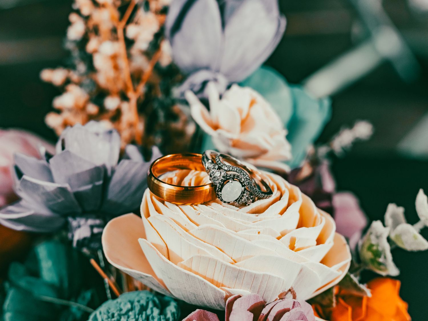 Wedding rings rest atop a delicate cream-colored rose surrounded by purple and peach flowers.