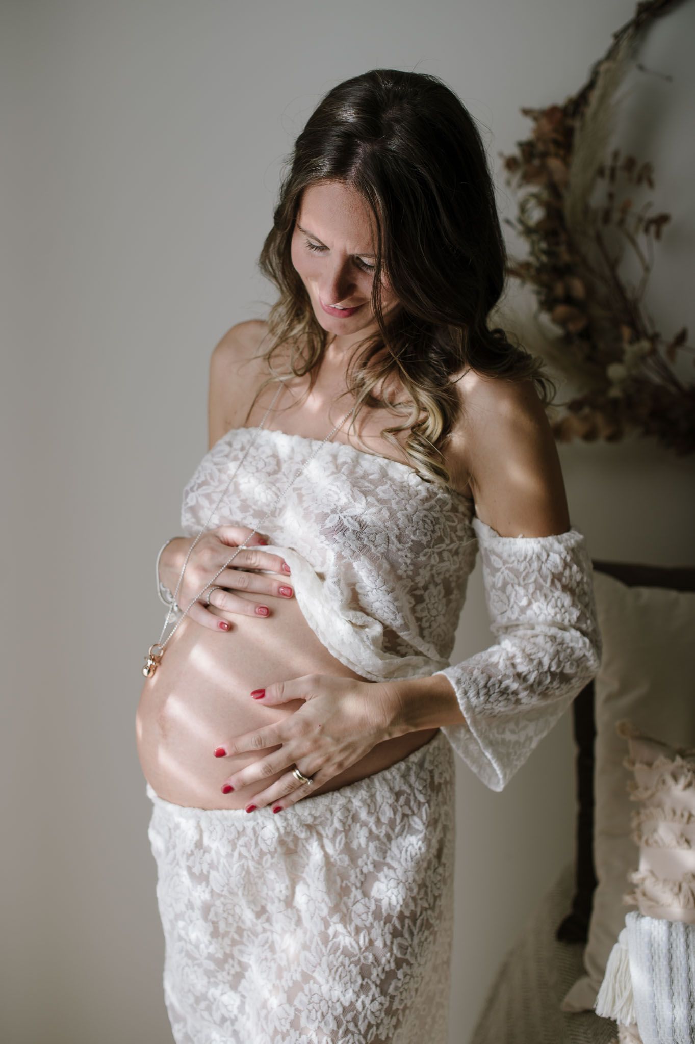 Expectant mother in white lace dress cradles her baby bump against a neutral background.